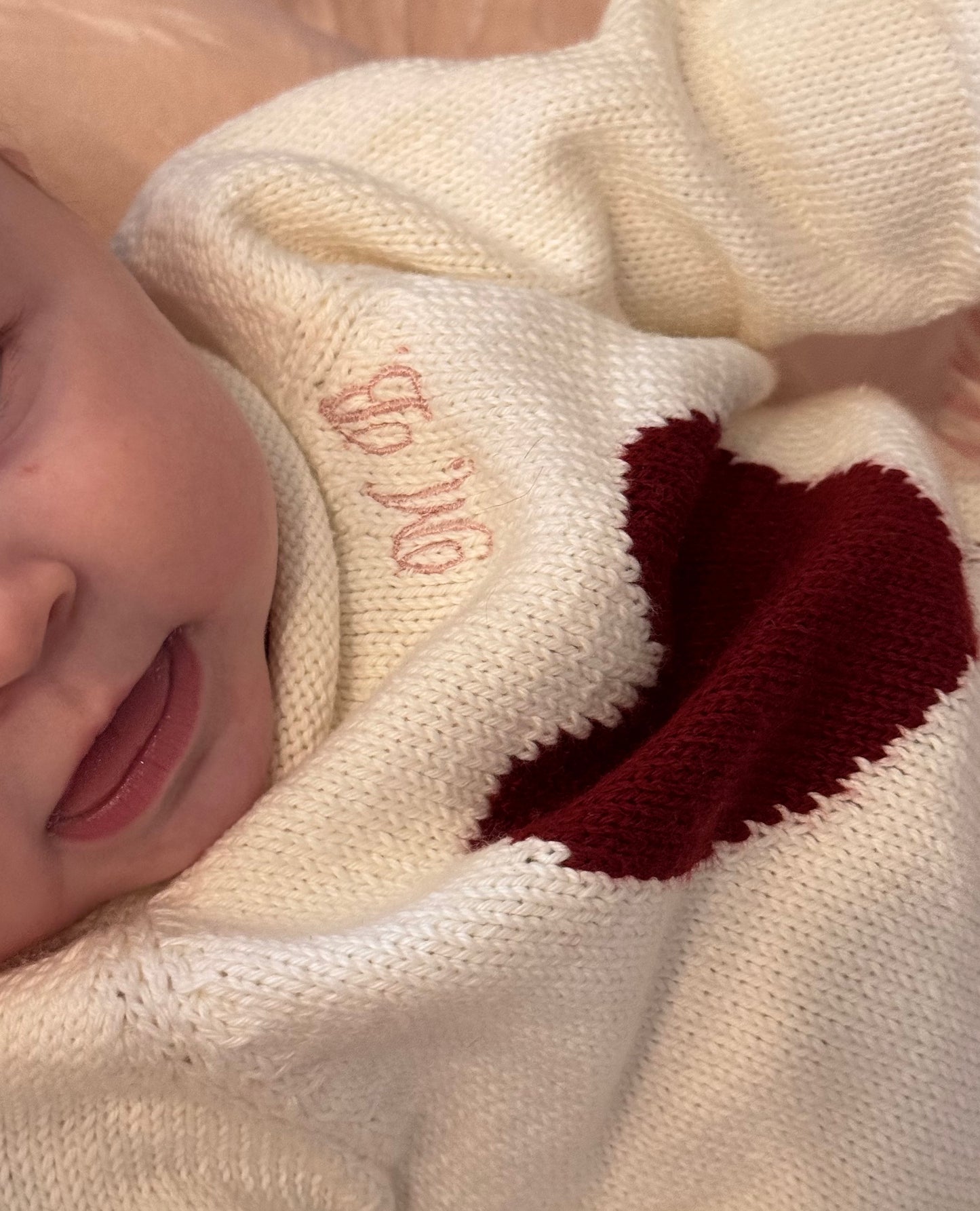 Close-up of a baby wrapped in a white sweater with red heart design, showing part of a name embroidered on the fabric.