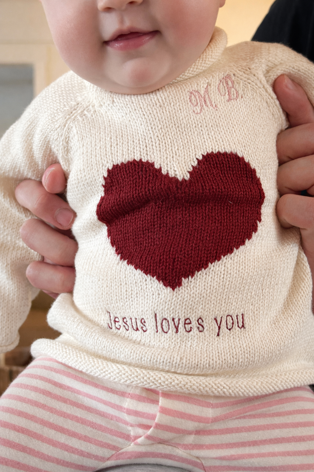 Baby wearing a sweater with a red heart and 'Jesus loves you' text, held by an adult.